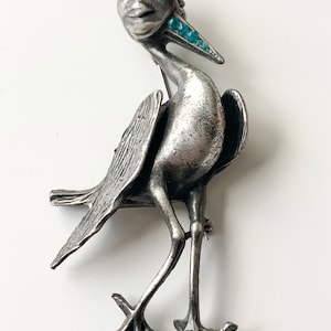 May include: A silver-toned bird brooch with blue rhinestone accents. The bird is standing on two legs with its wings spread out.