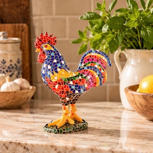 May include: A decorative rooster figurine crafted from colorful mosaic glass tiles. The bird features a vibrant mix of red, blue, yellow, and green shards, with a bright red comb and wattle, standing on a textured green mosaic base.