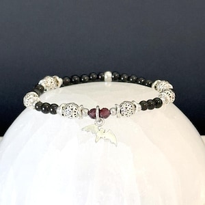May include: A black beaded bracelet with silver filigree beads and a small, dangling bat charm. The bracelet features a central dark red bead and is displayed on a white, rounded surface against a dark background. The bracelet is a gothic style.