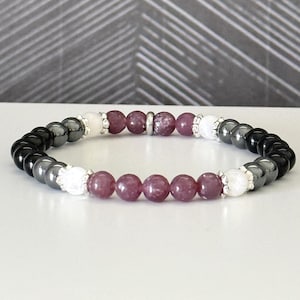 May include: A beaded stretch bracelet featuring a repeating pattern of polished black hematite beads, translucent white quartz beads, and deep mauve-colored round beads, accented with small silver-toned metal spacers throughout the circular design.