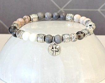 May include: A beaded stretch bracelet featuring a mix of grey, white, and speckled stone beads interspersed with silver-toned metal spacers. A circular silver-toned charm with an embossed cross hangs from the center of the bracelet.