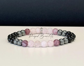 May include: A beaded bracelet featuring a mix of colors. The bracelet has black, gray, pink, and clear beads. The beads are arranged in a circular pattern. The text "Peaped Bracelets" is visible in the image.