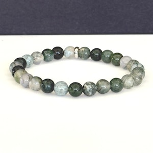 May include: A beaded bracelet featuring a variety of green and gray natural stone beads. The bracelet is strung on an elastic band and has a silver-colored spacer bead. The beads are approximately 0.3 inches in diameter.