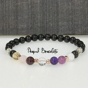 May include: A black beaded bracelet with a variety of colored gemstone beads, including rose quartz, garnet, clear quartz, amethyst, and pink amethyst. The bracelet is accented with silver and gold beads. The text "Peapod Bracelets" is visible on the bracelet.