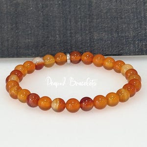 May include: An orange beaded bracelet with a silver clasp. The bracelet is made of small, round beads in shades of orange, red, and cream. The text "Peapod Bracelets" is visible in the image.