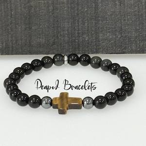 May include: Black beaded bracelet with a tiger's eye cross charm and hematite beads. The bracelet has a silver spacer bead and the text "Peapod Bracelets" is visible in the image.