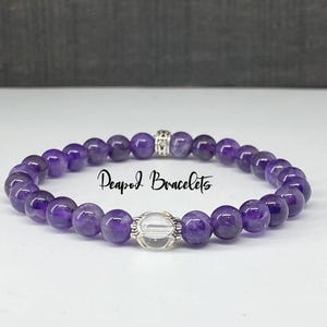 May include: A purple amethyst gemstone bracelet with a clear quartz bead charm and silver accents. The bracelet is made with round beads and has the text "Peapod Bracelets" on it.