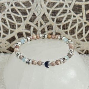 May include: A beaded bracelet featuring a mix of light brown, blue, and silver beads. The bracelet includes a small silver crescent moon charm. The beads are strung on a stretchy cord, creating a delicate and stylish accessory.