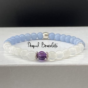 May include: A bracelet made with light blue and white beads with a single purple amethyst bead in the center. The bracelet has a silver clasp and the text "Peapod Bracelets" is visible in the image.