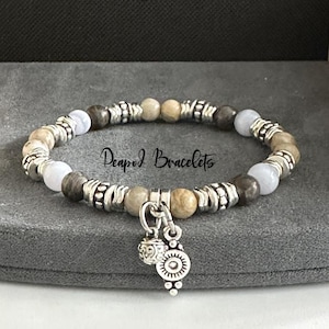 May include: A beaded bracelet with a silver charm. The bracelet features light brown, gray, and white beads with silver spacers. The charm is a silver circle with a smaller silver ball attached.