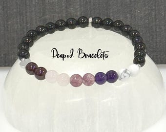 May include: A black beaded bracelet with a silver charm and a row of colorful gemstone beads. The beads are red, pink, light pink, purple, and white. The bracelet is on a white surface. "Deapod Bracelets" is written on the surface.