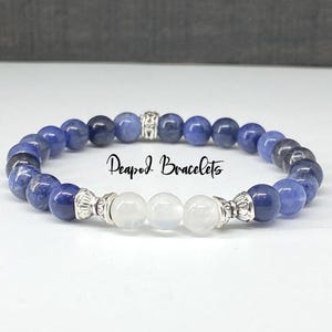 May include: A beaded bracelet featuring dark blue and white beads. The bracelet has a silver-colored accent piece and three white beads in the center. The text "Deapod Bracelets" is written in cursive across the image.