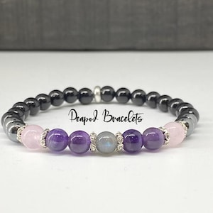 May include: A beaded bracelet with black, purple, pink, and gray stones. The bracelet has a silver clasp and the text "Peapod Bracelets" is visible.