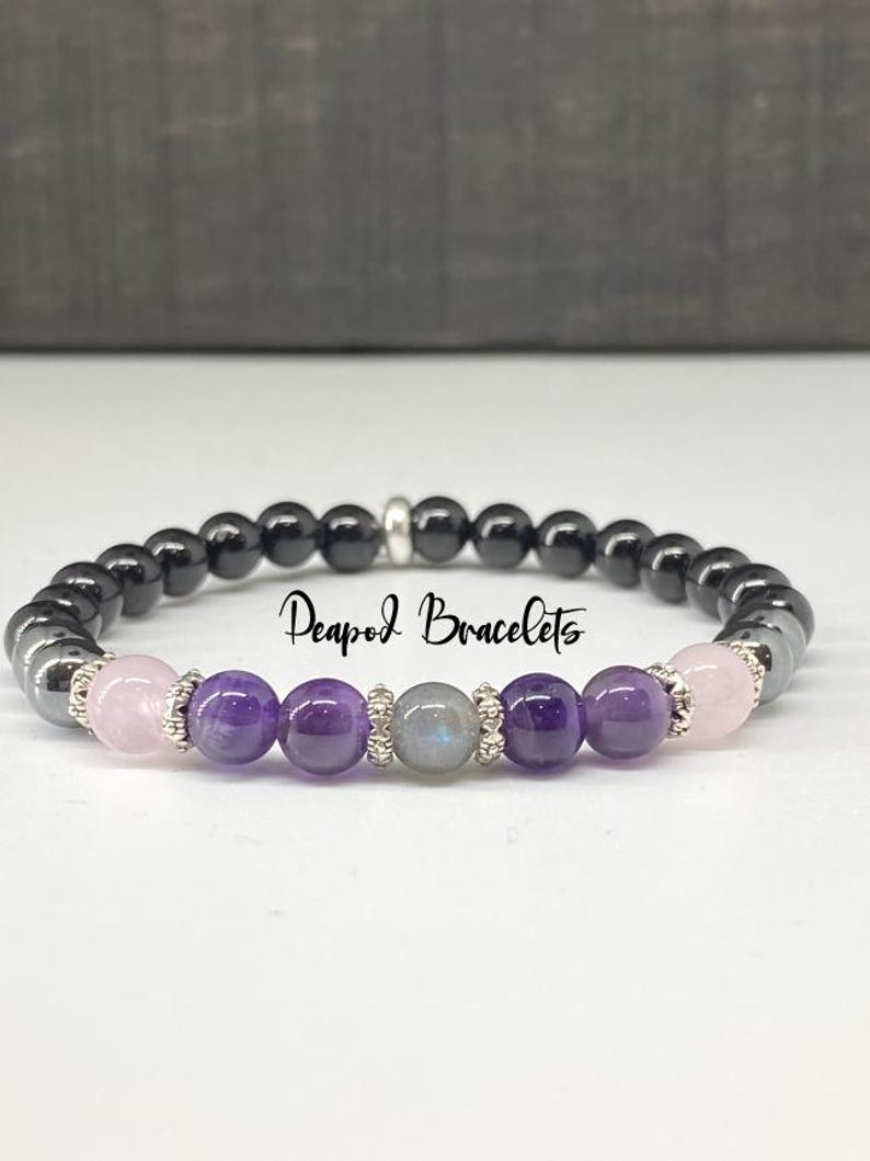 May include: A beaded bracelet with black, purple, pink, and gray stones. The bracelet has a silver clasp and the text "Peapod Bracelets" is visible.