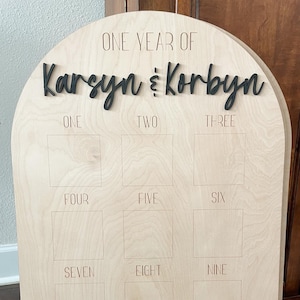 May include: A wooden milestone board with an arched top. The words "ONE YEAR OF Karsyn & Korbyn" are in black script. Below are twelve squares labeled with the numbers one through twelve. The board is made of light-colored wood.
