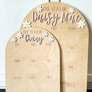 May include: Two arched wooden milestone boards for a first birthday. The larger board displays 'One Year of Daisy Mae' with pink lettering and white daisy accents.  The smaller board shows numbers one through twelve in a grid layout. Both boards feature engraved numbers and daisy embellishments. The boards are light wood tone.
