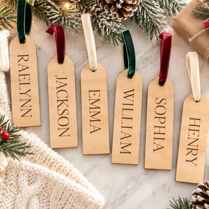 May include: Six rectangular wooden tags with rounded tops, each engraved with a name: RAELYNN, JACKSON, EMMA, WILLIAM, SOPHIA, and HENRY. Each tag features a velvet ribbon loop in either dark green, deep red, or cream, hanging for display.