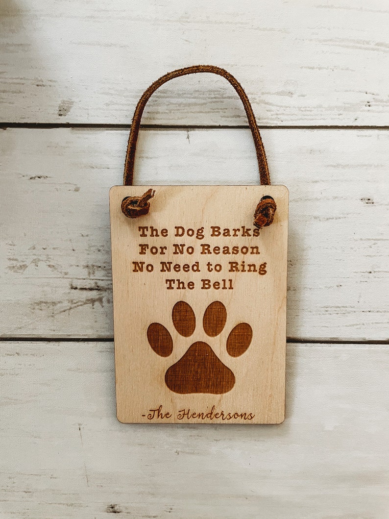 May include: A rectangular wooden sign with a leather loop for hanging. The sign features the text "The Dog Barks For No Reason No Need to Ring The Bell" and a paw print. The name "-The Hendersons" is below the paw print.