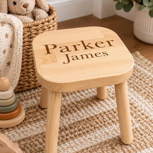 May include: A small, light-colored wooden stool with four rounded legs and a square seat. The top of the seat features the engraved text 'Parker James' in a dark, serif font. The stool has a smooth, natural wood grain finish throughout.