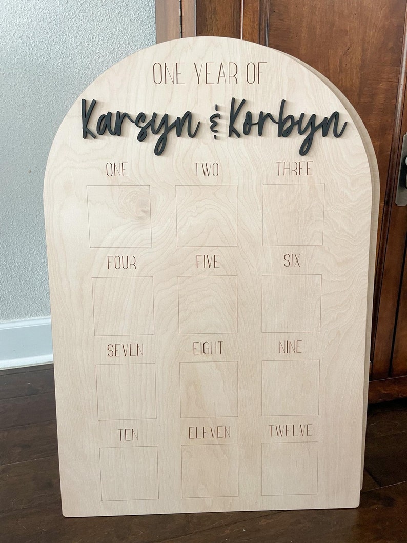 May include: A wooden milestone board with an arched top. The words "ONE YEAR OF Karsyn & Korbyn" are in black script. Below are twelve squares labeled with the numbers one through twelve. The board is made of light-colored wood.