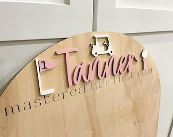May include: A light wood sign with a pink and white golf theme. The sign says "Tanner mastered her first year" with a golf cart, golf flag, and golf ball.