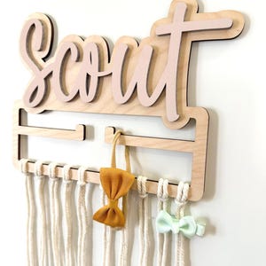 May include: Wooden hair bow holder with the name "Scout" in pink script. The holder has a macrame design with hanging cords and displays two bows, one yellow and one mint green.