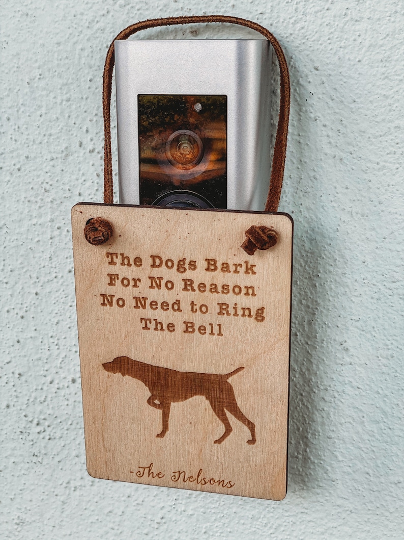 May include: A wooden sign with the text 'The Dogs Bark For No Reason. No Need to Ring The Bell - The Nelsons' and a silhouette of a dog. The sign is hanging on a wall with a silver doorbell camera above it.