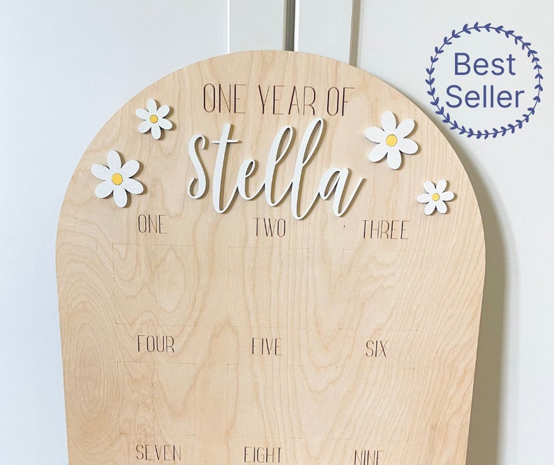 Puede incluir: Un letrero de madera en forma de arco con el texto "ONE YEAR OF Stella" y los n&uacute;meros del uno al nueve. El letrero est&aacute; decorado con flores de margarita blancas con centros amarillos.