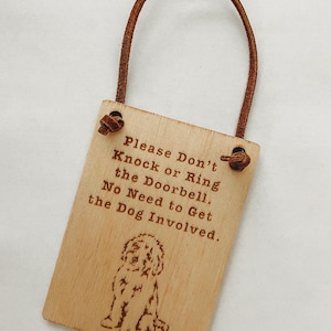 May include: A rectangular wooden sign with a brown leather loop. The sign has engraved text that reads "Please Don't Knock or Ring the Doorbell. No Need to Get the Dog Involved." A dog illustration is below the text.