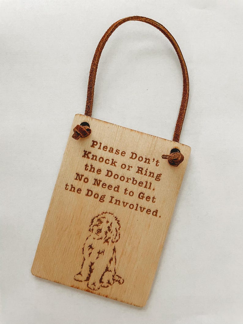 May include: A rectangular wooden sign with a brown leather loop. The sign has engraved text that reads "Please Don't Knock or Ring the Doorbell. No Need to Get the Dog Involved." A dog illustration is below the text.