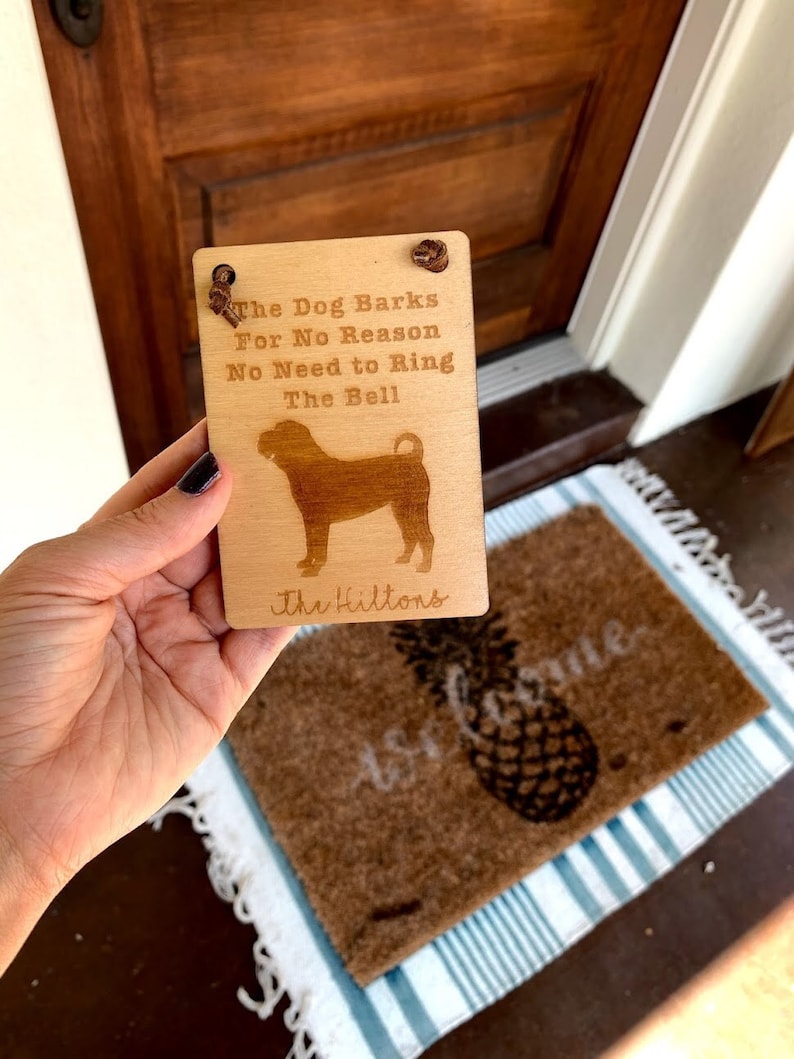 May include: Wooden sign with the text "The Dog Barks For No Reason No Need to Ring The Bell" and a dog silhouette. The sign also includes the family name "The Hiltons". The sign is held in front of a doormat.
