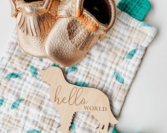 Puede incluir: Un par de mocasines dorados de cuero para bebé con flecos descansa sobre una manta de muselina con un estampado de hojas. También hay un letrero de madera con forma de perro con las palabras "hello world". La manta tiene detalles turquesa.