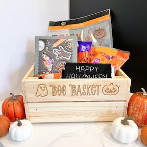 May include: A wooden crate with the words "Boo Basket" and a ghost and pumpkin face carved into the wood. The crate is filled with Halloween treats, including candy, cookies, and a black cloth with the words "Happy Halloween" embroidered on it.