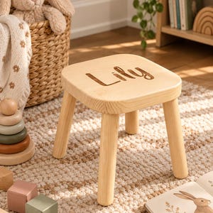 May include: A small, light-colored wooden stool with four rounded legs and a square seat. The name Lily is engraved in a cursive script on the top surface of the seat. The stool is constructed from natural wood with a visible grain pattern throughout.