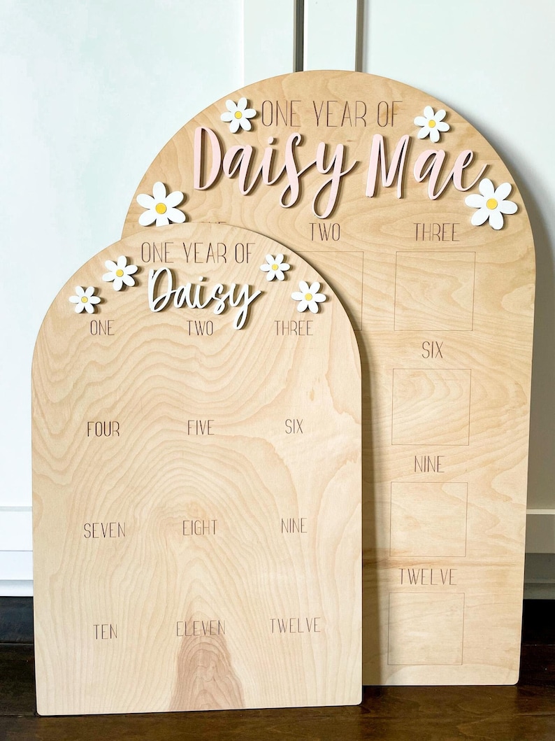 May include: Two arched wooden milestone boards for a first birthday. The larger board displays 'One Year of Daisy Mae' with pink lettering and white daisy accents.  The smaller board shows numbers one through twelve in a grid layout. Both boards feature engraved numbers and daisy embellishments. The boards are light wood tone.