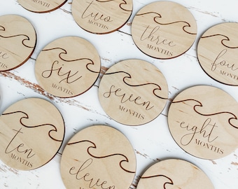 May include: A set of 11 wooden baby milestone discs with wave designs and engraved text. The discs feature the phrases "hello world", "one month", "two months", "three months", "four months", "six months", "seven months", "eight months", "ten months", "eleven months", and "one year".