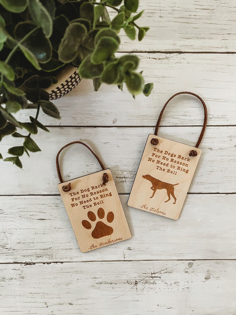 May include: Two rectangular wooden signs with brown leather loops. One sign features a paw print, the other a dog silhouette. Both signs have the text "The Dog Barks For No Reason No Need to Ring The Bell" and a family name.