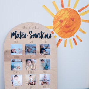 Pode incluir: Uma placa de madeira com o topo arredondado, com as palavras "One Year of Mateo Santino" em letras pretas. A placa &eacute; dividida em doze quadrados, cada um contendo uma foto de um menino beb&ecirc; em diferentes idades, etiquetados com o m&ecirc;s correspondente.