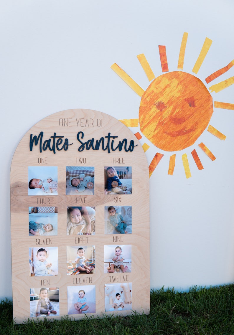 Pode incluir: Uma placa de madeira com o topo arredondado, com as palavras "One Year of Mateo Santino" em letras pretas. A placa &eacute; dividida em doze quadrados, cada um contendo uma foto de um menino beb&ecirc; em diferentes idades, etiquetados com o m&ecirc;s correspondente.