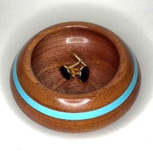 Puede incluir: Un cuenco de madera con una banda azul claro alrededor del borde. El cuenco está hecho de madera oscura y tiene un acabado liso y pulido. Hay dos gemelos dentro del cuenco.