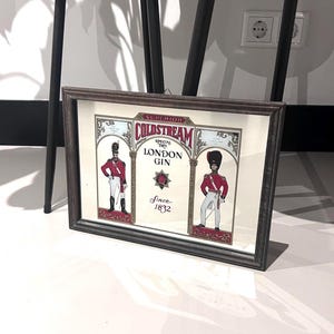 May include: A framed print featuring a vintage advertisement for Coldstream London Gin. The print showcases two soldiers in red coats and bearskin hats, with the text "Superior Coldstream London Gin Since 1832."