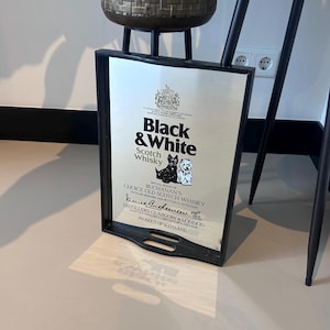 Op de afbeelding: Een zwart omrand bord met het "Black & White" Scotch Whisky-logo. Het logo bevat de merknaam, een wapen en illustraties van een zwarte en een witte hond. Het bord bevat ook de tekst "Buchanan's Choice Old Scotch Whisky".