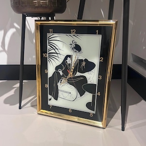 Könnte beinhalten: Eine rechteckige Uhr mit goldenem Rahmen und schwarzem Zifferblatt. Das Zifferblatt zeigt eine schwarz-weiße Art-Déco-Illustration einer Frau mit Hut. Goldene Ziffern markieren die Stunden. Die Uhr ist an einer weißen Wand aufgestellt.