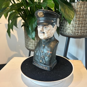 May include: A cast iron bust of a soldier wearing a military uniform and a cap. The bust is painted in shades of green, black, and white. The bust is inscribed with the name "PERSHING".