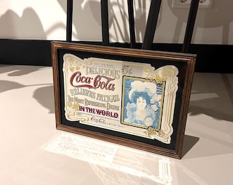 May include: A rectangular framed Coca-Cola advertisement sign featuring a vintage illustration of a person in a blue hat. The text reads: Delicious Coca-Cola, Relieves Fatigue, The Most Refreshing Drink In The World, Sold Everywhere In Glass Or Bottle.