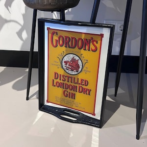 May include: A framed sign for Gordon's Distilled London Dry Gin. The sign features a yellow background with red and black text, including the brand name and product details. The frame is black.