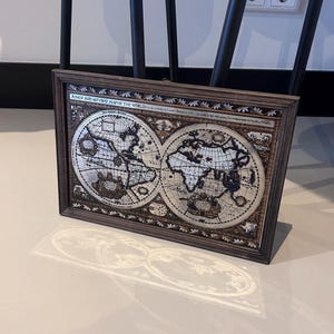 Op de afbeelding: Een ingelijste vintage wereldkaart. De kaart staat in een donkerhouten lijst en toont een gedetailleerde weergave van de continenten en oceanen. De kaart heeft een sepia-tint met ingewikkelde details en tekst.