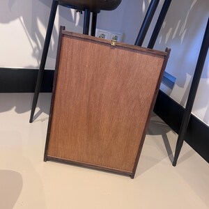 K&ouml;nnte beinhalten: Ein rechteckiger Holzrahmen mit hellbrauner Oberfl&auml;che. Der Rahmen hat einen dunkelbraunen Rand und einen kleinen goldfarbenen Verschluss. Der Holzrahmen lehnt an einer Wand.