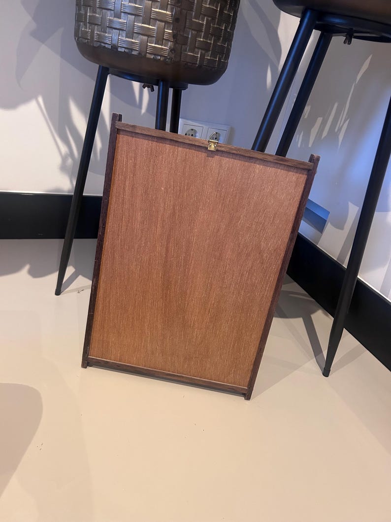 K&ouml;nnte beinhalten: Ein rechteckiger Holzrahmen mit hellbrauner Oberfl&auml;che. Der Rahmen hat einen dunkelbraunen Rand und einen kleinen goldfarbenen Verschluss. Der Holzrahmen lehnt an einer Wand.