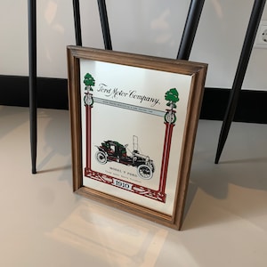 May include: A vintage framed mirror with a Ford Motor Company advertisement featuring a black and white illustration of a Model T Ford car. The text reads "Ford Motor Company, Low Priced Quality is a Low Priced Car, Model T Ford, Three-seater, $575, November 1, 1910".